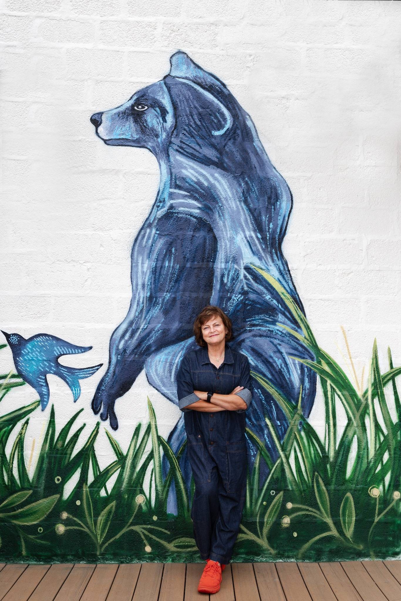 Woman standing in front of a mural of a blue bear and bird on a white brick wall.